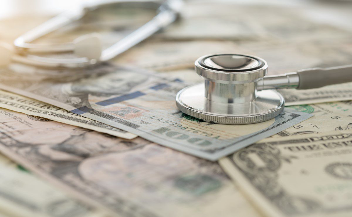 A stethoscope rests on top of various US dollar bills, representing the concept of healthcare costs or medical expenses.