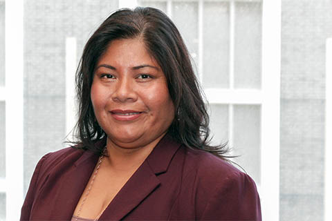 A woman with dark hair wearing a maroon blazer smiles in front of a window with a grid pattern.