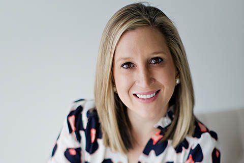 A woman with straight blonde hair and a patterned blouse smiles at the camera against a plain light background.