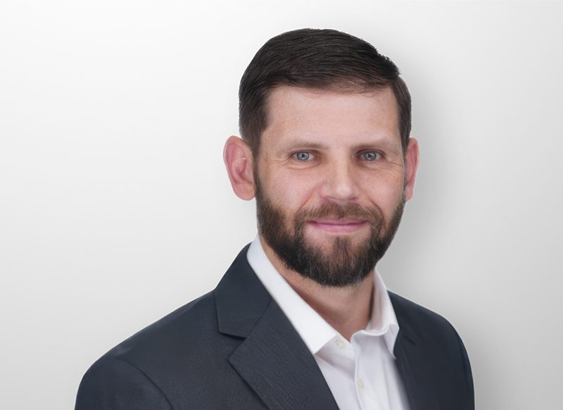 A man with short dark hair and a beard wearing a dark suit jacket and white shirt, posing against a plain light background.