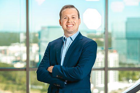 A man in a blue suit stands with arms crossed, smiling at the camera, in front of large windows overlooking a cityscape.