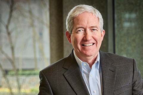 A man with short gray hair in a dark suit jacket and light blue shirt smiles while standing indoors near a window.