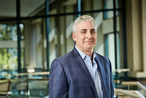 A man with gray hair wearing a blue plaid blazer and light blue shirt stands indoors in front of large windows.