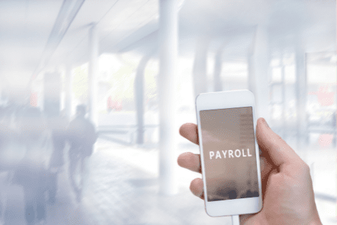 A hand holds a smartphone displaying the word PAYROLL on the screen, with a blurred modern office background and people walking.