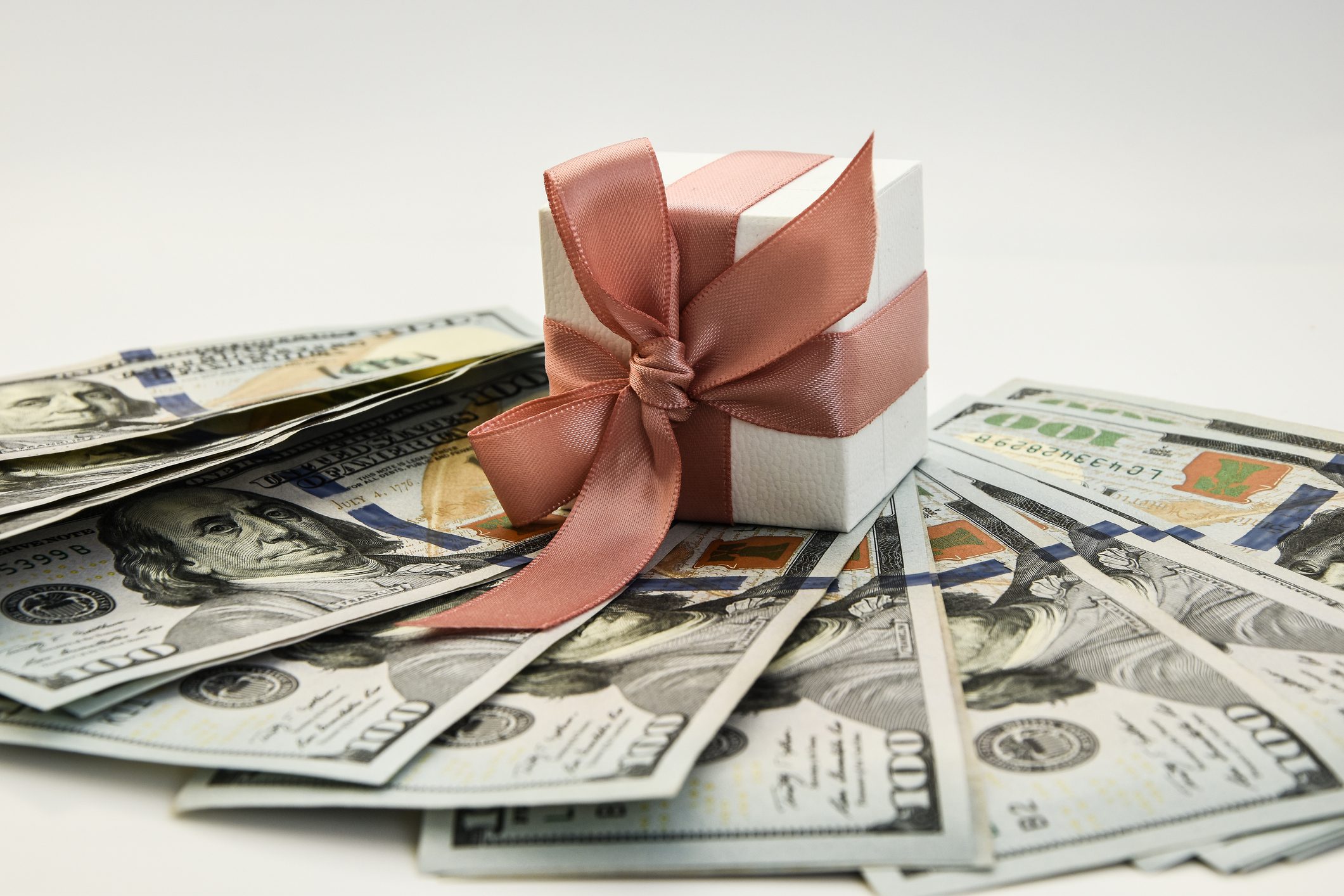 A small white gift box with a pink ribbon bow sits on top of scattered US $100 bills against a plain white background.