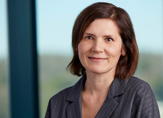 A woman with short brown hair wearing a gray pinstripe blazer stands indoors in front of a window with a blurred outdoor background.