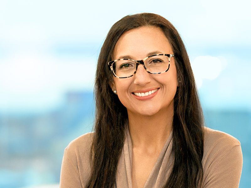 A woman with long dark hair and glasses smiles at the camera while wearing a beige top, with a blurred blue background.