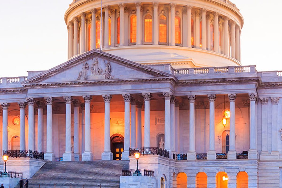 The front facade of the United States Capitol building at dusk, with illuminated windows and columns visible.