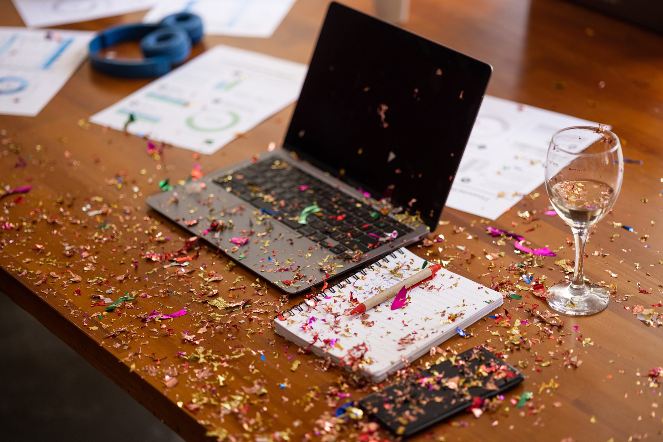A laptop, notebook, and wine glass sit on a wooden table covered in colorful confetti, with scattered papers and headphones in the background.