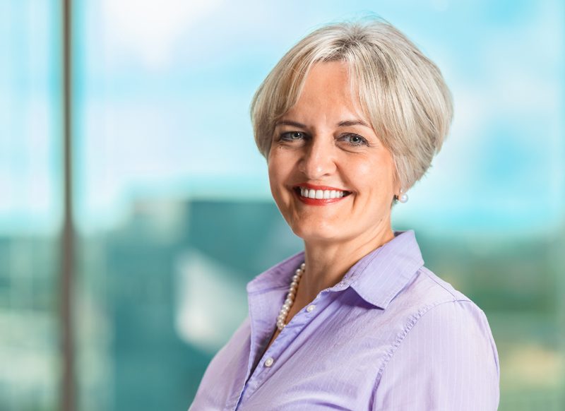Smiling older woman with short gray hair wearing a light purple blouse and pearl necklace, standing indoors with a blurred window background.