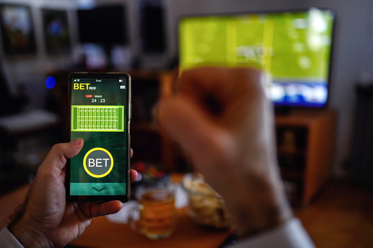 A person holds a smartphone displaying a sports betting app, with a football game on TV in the background and their fist raised in apparent excitement.