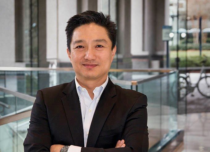 A man in a dark suit stands indoors with his arms crossed, smiling at the camera. The background features glass railings and large windows.