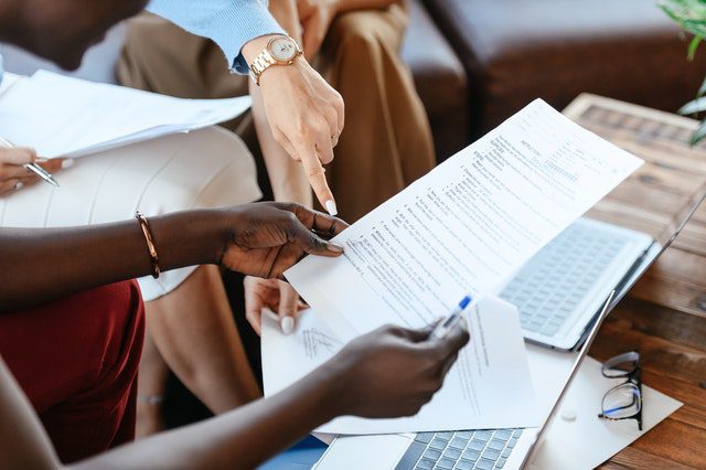 Two people sitting at a table reviewing a printed document together, with laptops and papers nearby. One person is pointing at the document.