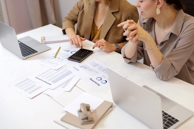 Two people sit at a desk with laptops, papers, and a calculator, reviewing financial documents and charts.