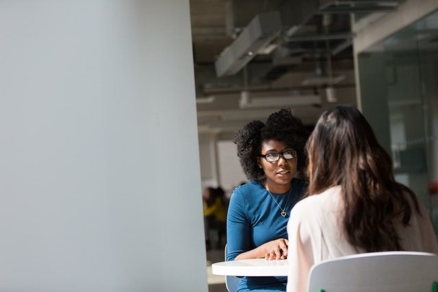 Two women sit across from each other at a table in a bright, modern office, engaged in a conversation.