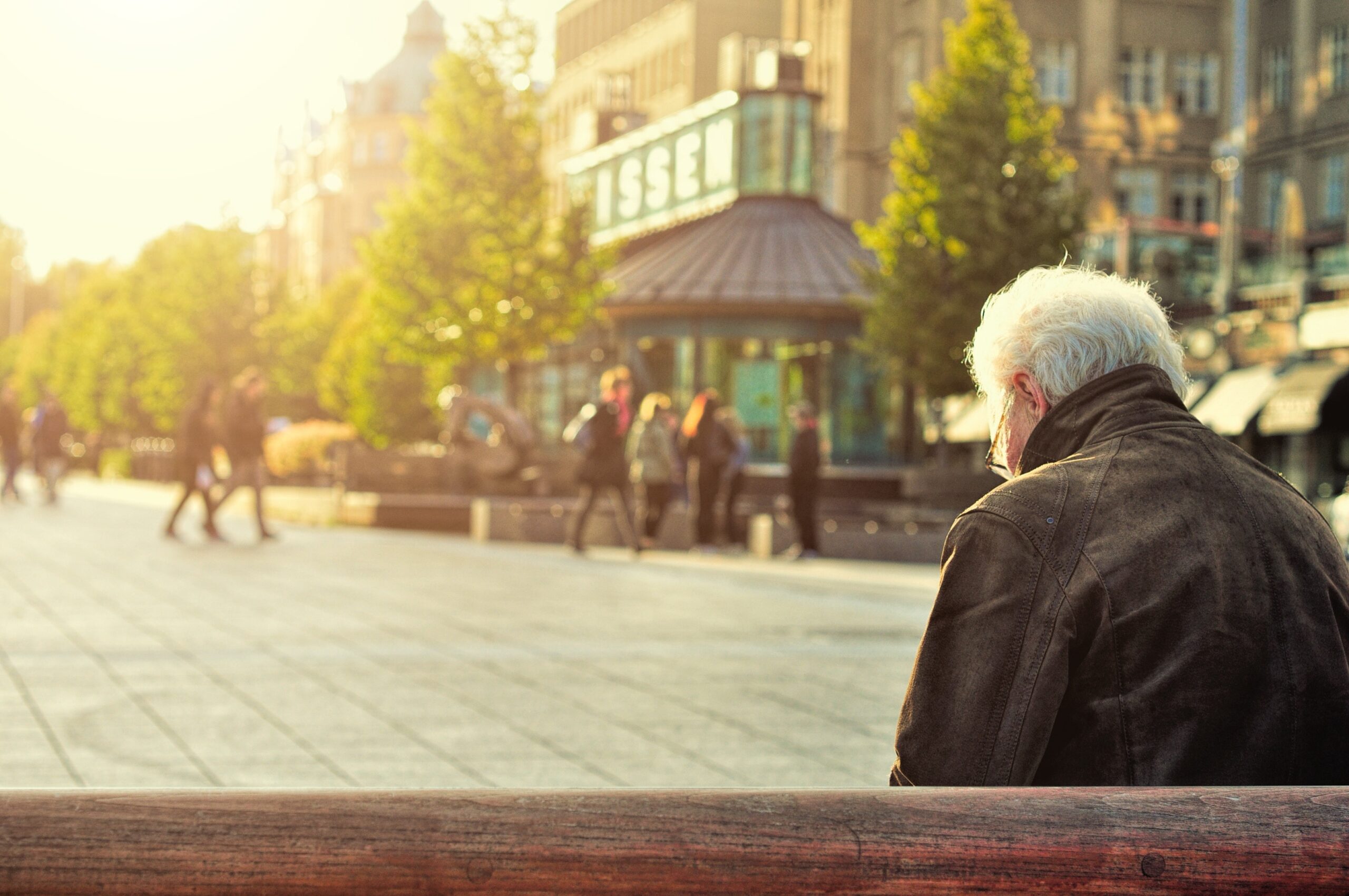 An older man with white hair sits alone on a bench in an urban square, while people walk by in the background on a sunny day.