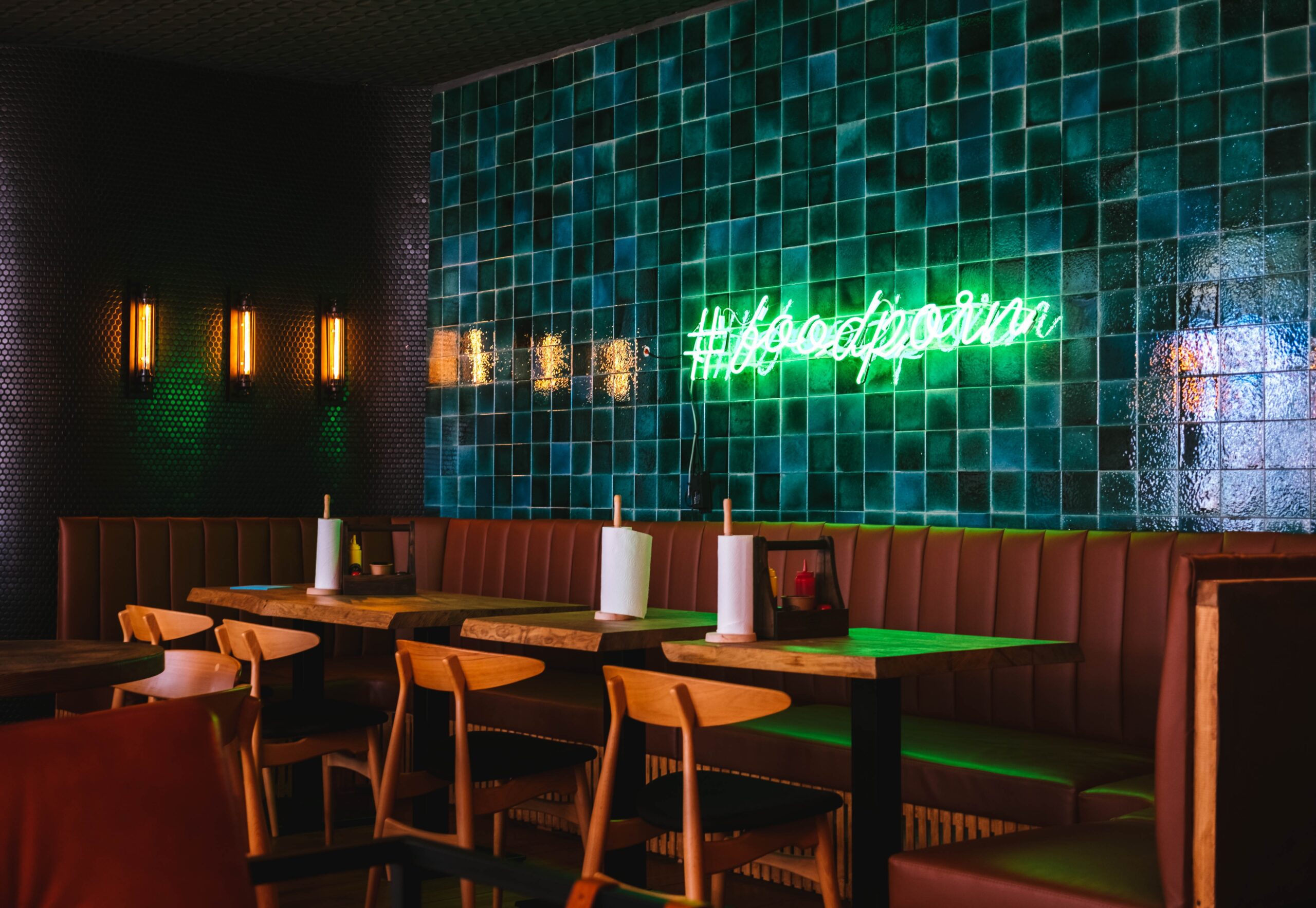 A modern restaurant interior with wooden tables and chairs, brown leather seating, green tiled wall, and a neon sign reading #foodporn.