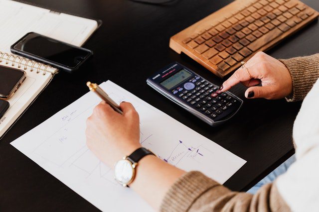 Person writing on a paper and using a calculator at a desk with a keyboard, notebook, and smartphone.