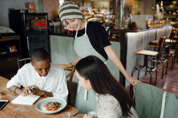A server in an apron stands by a table, smiling at two seated customers as they look at a plate of food in a restaurant setting.