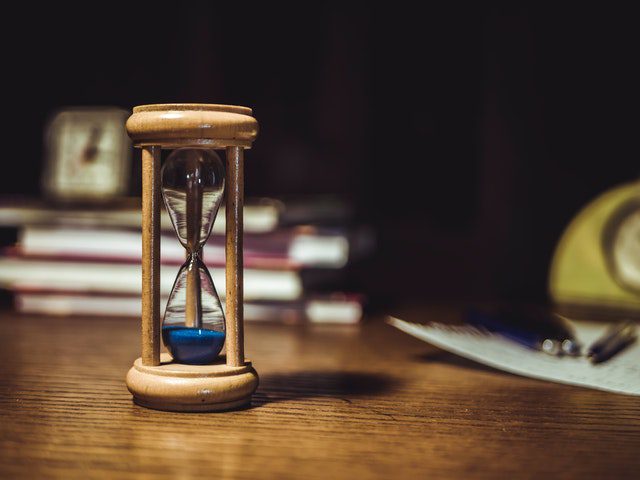 A wooden hourglass with blue sand sits on a wooden table, with blurred books, a clock, and papers in the background.