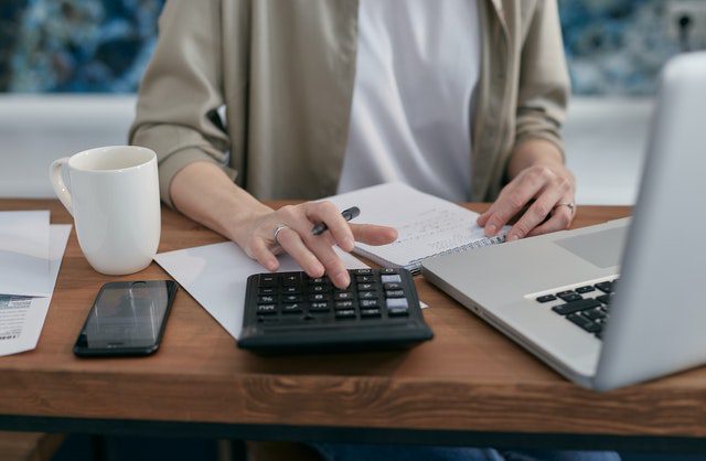 Person using a calculator at a desk with a notebook, smartphone, coffee mug, and laptop.