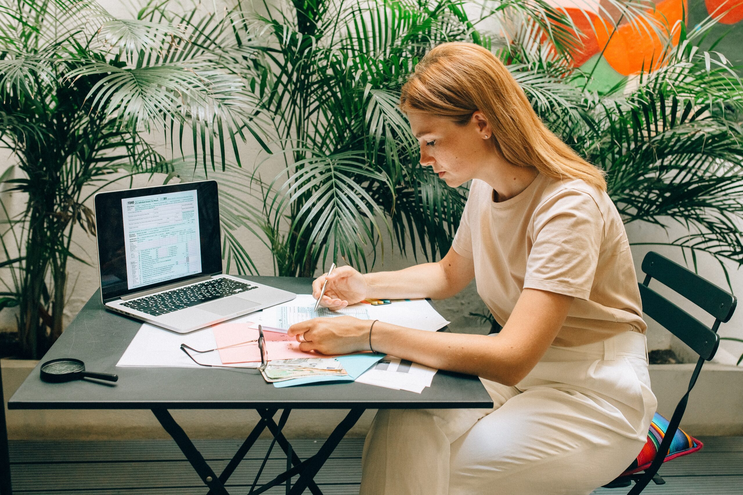 A woman sits at a black table working with papers and a laptop, surrounded by plants. She appears focused on writing or reviewing documents.