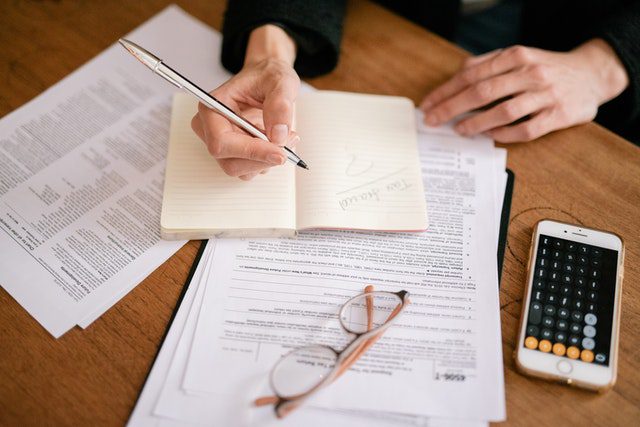 Person writing in a notebook with documents, glasses, and a smartphone displaying a calculator on a wooden table.