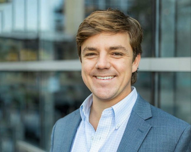 A man with light brown hair wearing a blue suit jacket and light blue shirt stands smiling in front of a glass building.