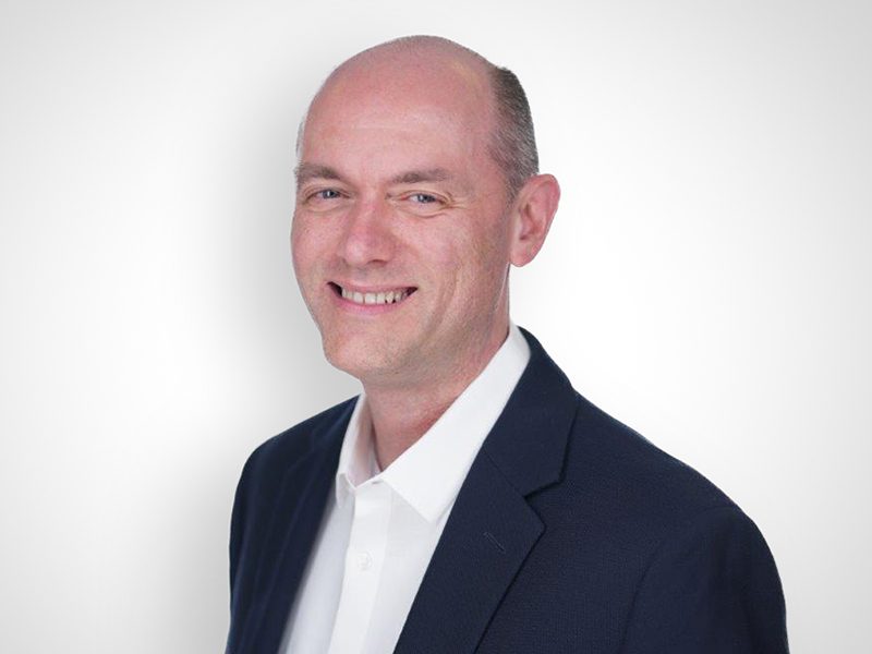 A man in a dark suit jacket and white shirt smiles at the camera against a plain white background.