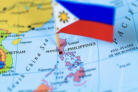 A small Philippine flag pinned on a map highlights Manila, the capital city, with neighboring countries and bodies of water also visible.