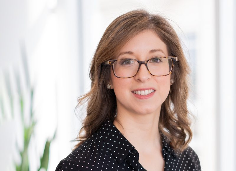A woman with shoulder-length brown hair and glasses, wearing a black blouse with white dots, stands indoors with a blurred background.