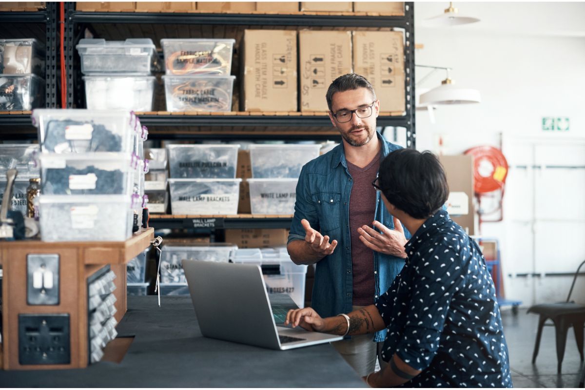 Two people having a discussion in a warehouse or storage area, with boxes and bins on shelves and a laptop on the counter.