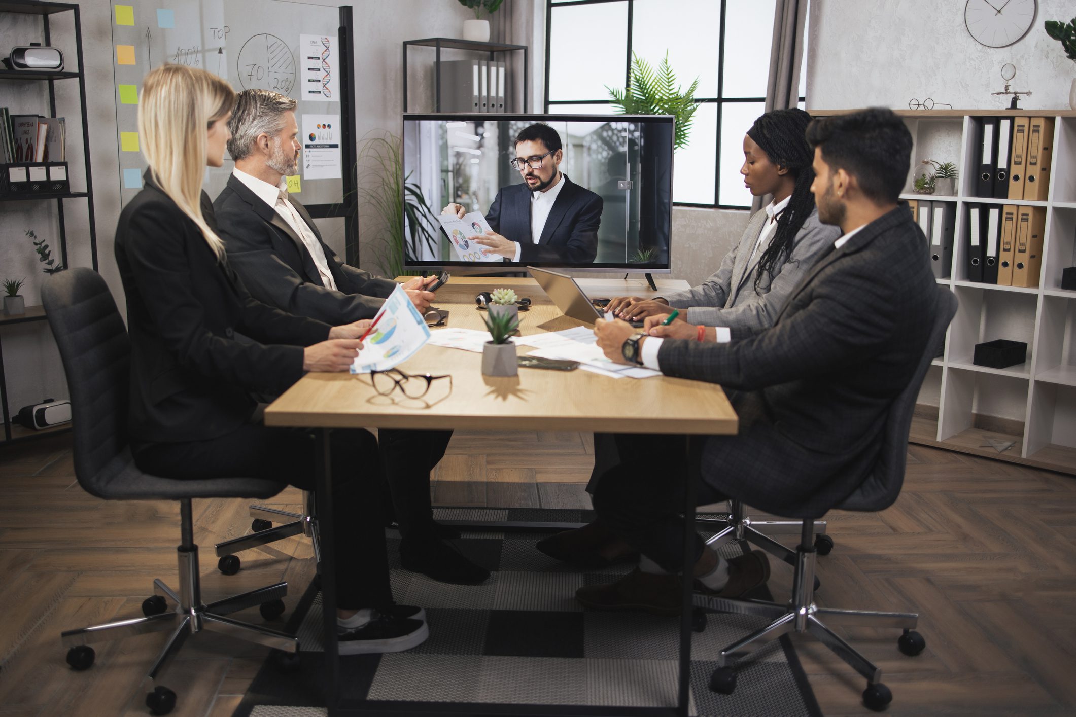Four people in business attire sit at a conference table having a video call with a man on a screen, surrounded by office supplies and charts.