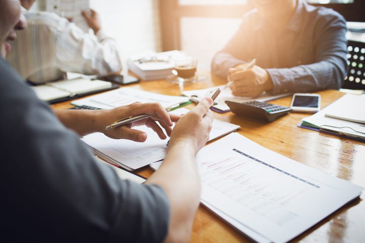 Three people sit at a table with documents, a calculator, and a smartphone, discussing paperwork in a bright office setting.