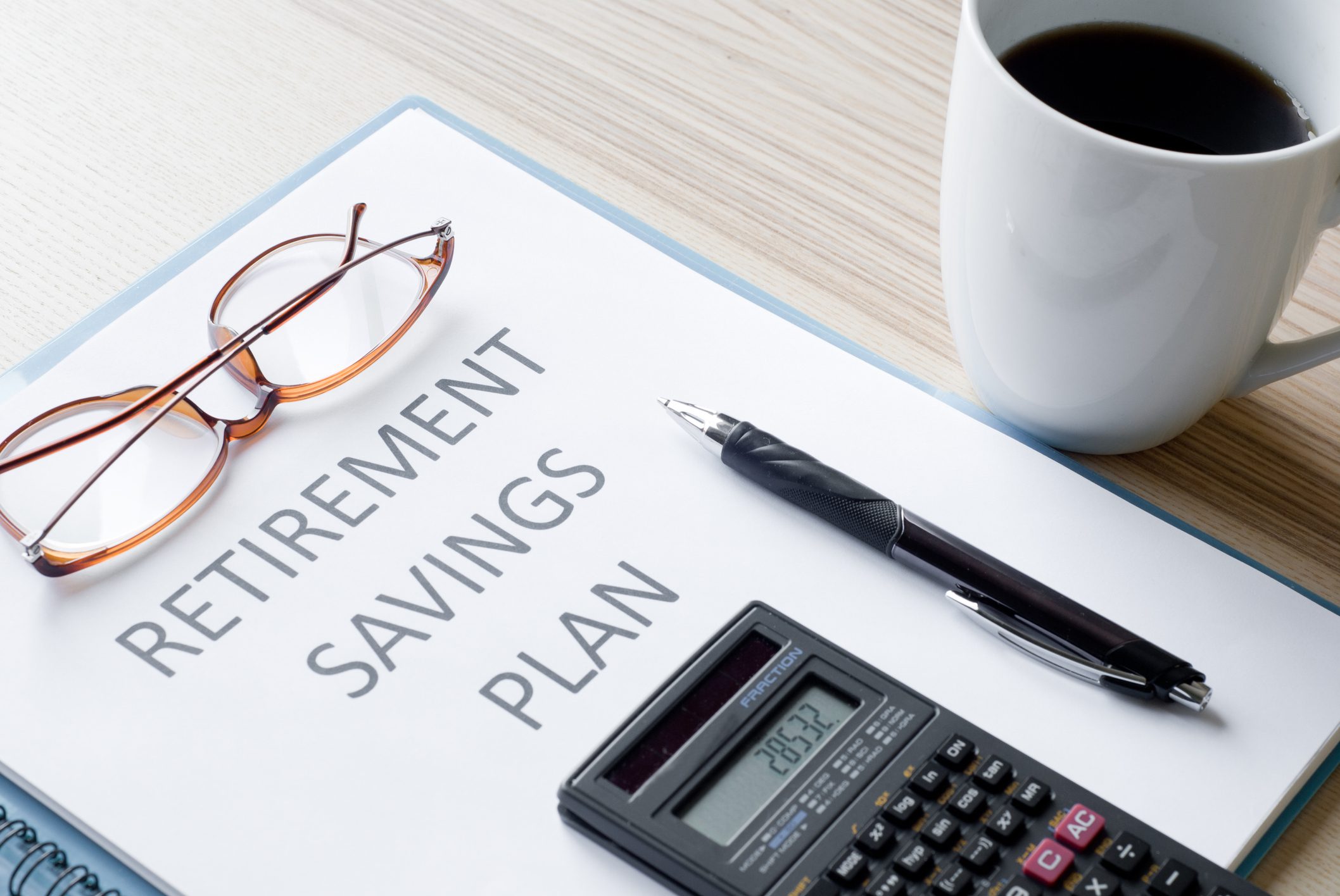 A document labeled Retirement Savings Plan on a desk with eyeglasses, a pen, a calculator, and a cup of coffee.