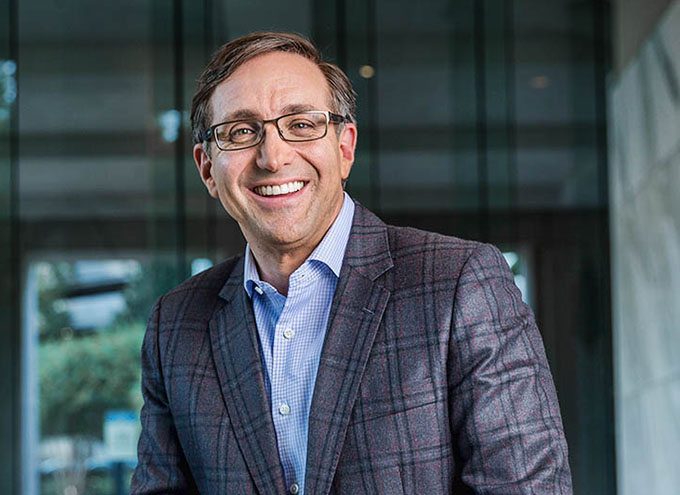A man wearing glasses, a blue checkered shirt, and a patterned blazer smiles while standing indoors in front of glass panels.