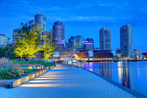 A waterfront cityscape at dusk with modern skyscrapers, illuminated trees, and a paved walkway along the water.
