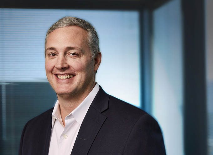 A man with short gray hair in a dark suit jacket and white shirt smiles while standing in an office with large windows.