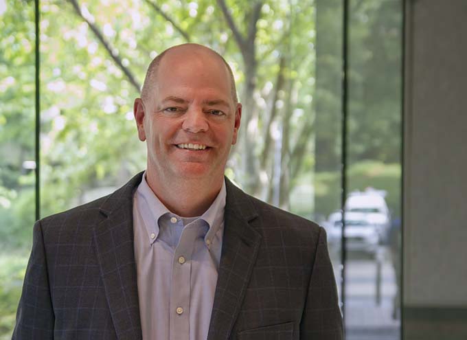 A man in a suit jacket and collared shirt stands indoors in front of large windows with trees and a parked car visible outside.