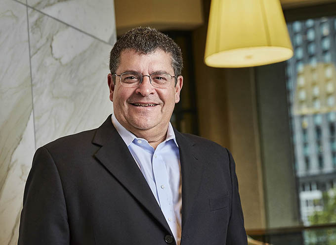 A man wearing glasses and a dark blazer smiles while standing indoors near a marble wall and large window with a city view.