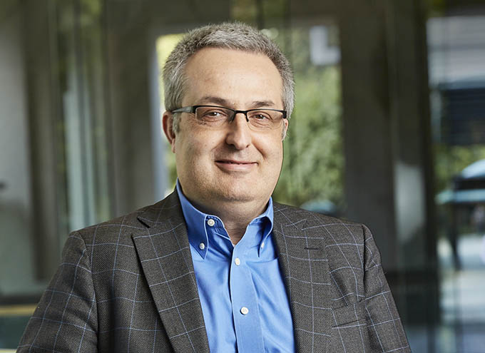 A middle-aged man with short gray hair and glasses, wearing a blue shirt and checkered blazer, is seated indoors with a blurred background.