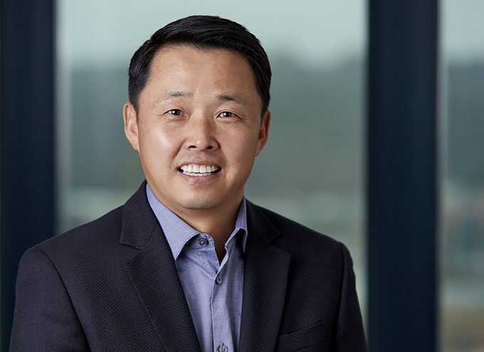 A man in a dark suit jacket and light blue shirt smiles at the camera, standing in front of large windows with a blurred outdoor background.