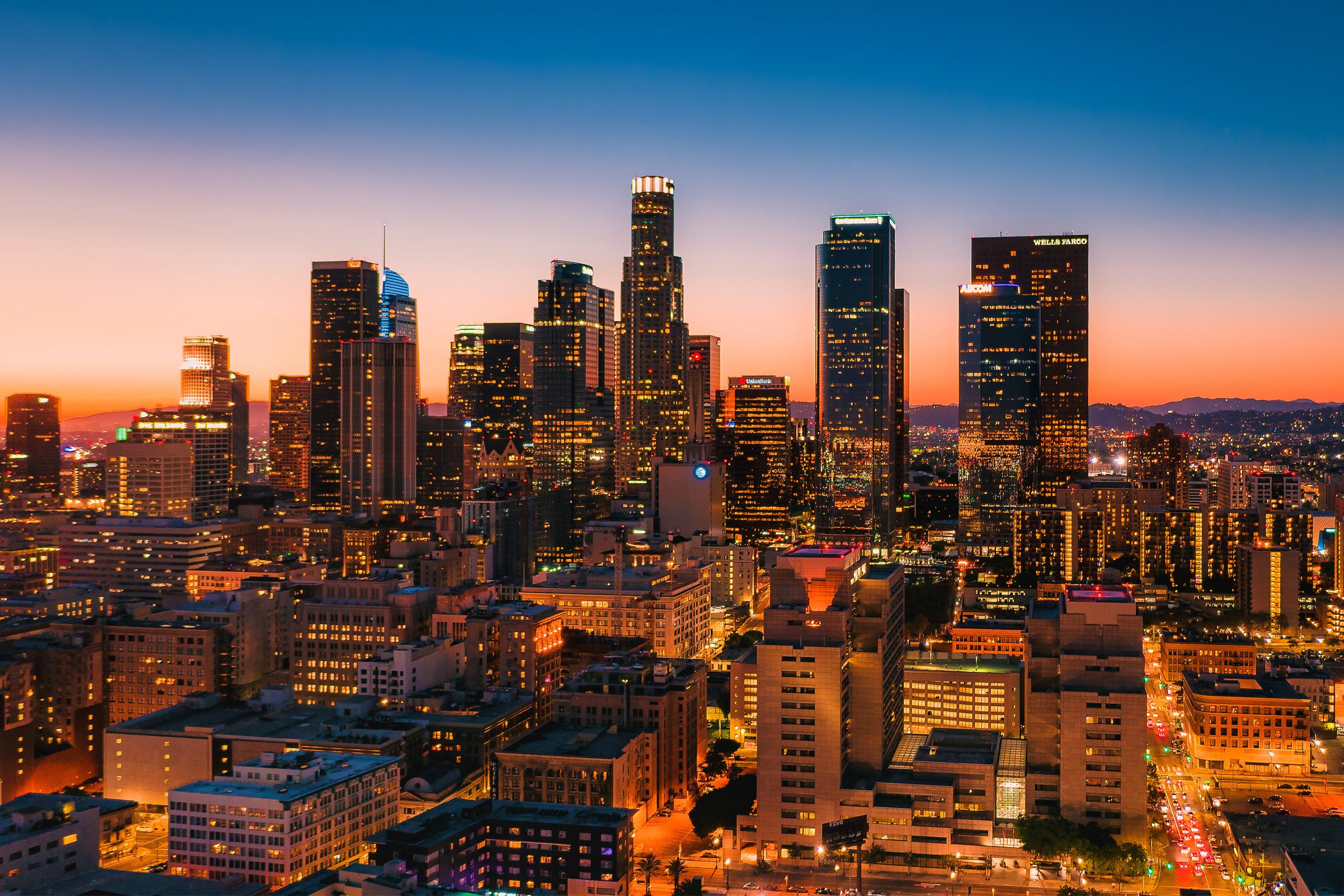 Downtown cityscape view of tall skyscrapers and buildings at sunset, with illuminated windows and a colorful twilight sky.