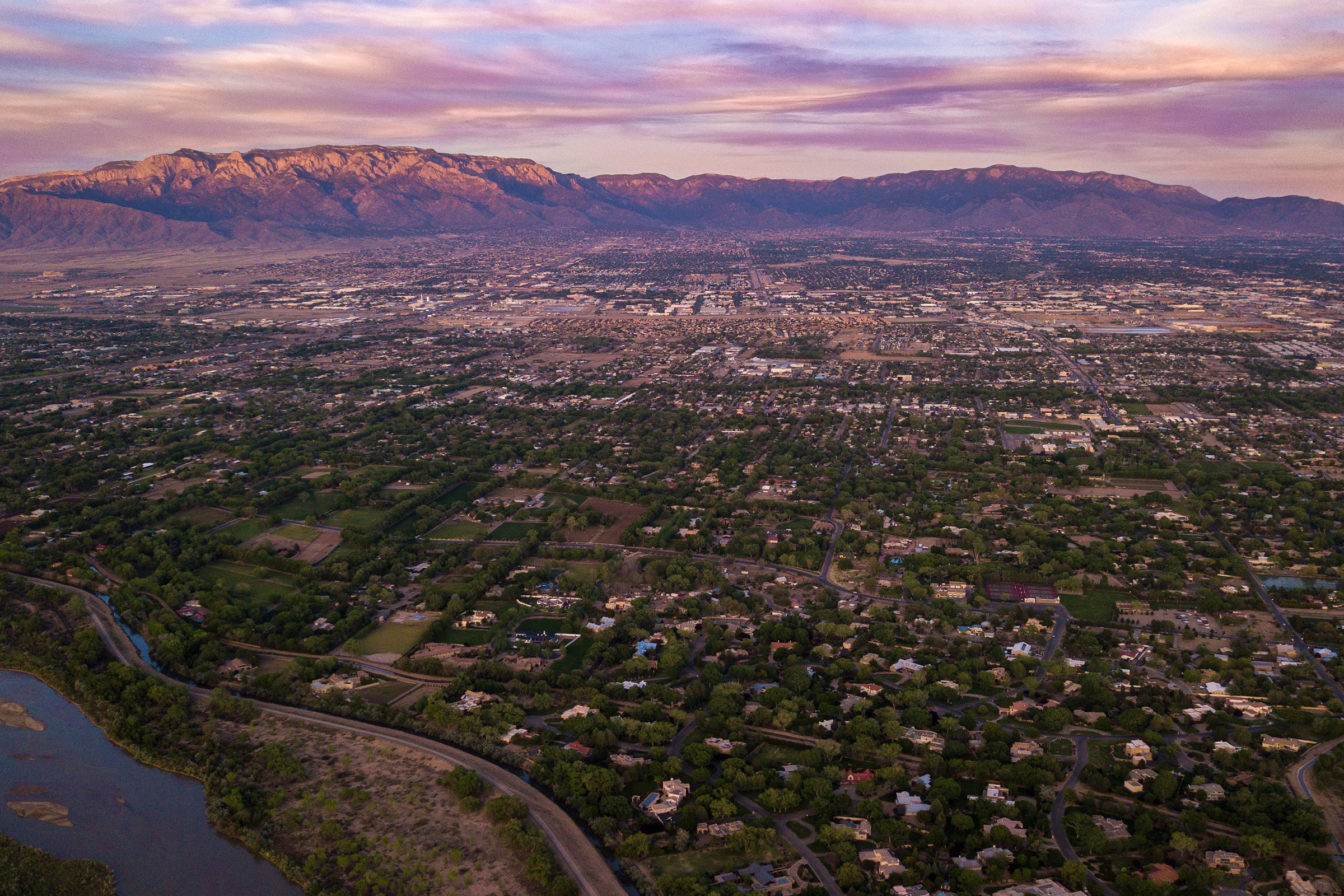 Aerial view of a sprawling city with scattered homes and greenery, bordered by mountains in the background under a colorful, cloudy sky.