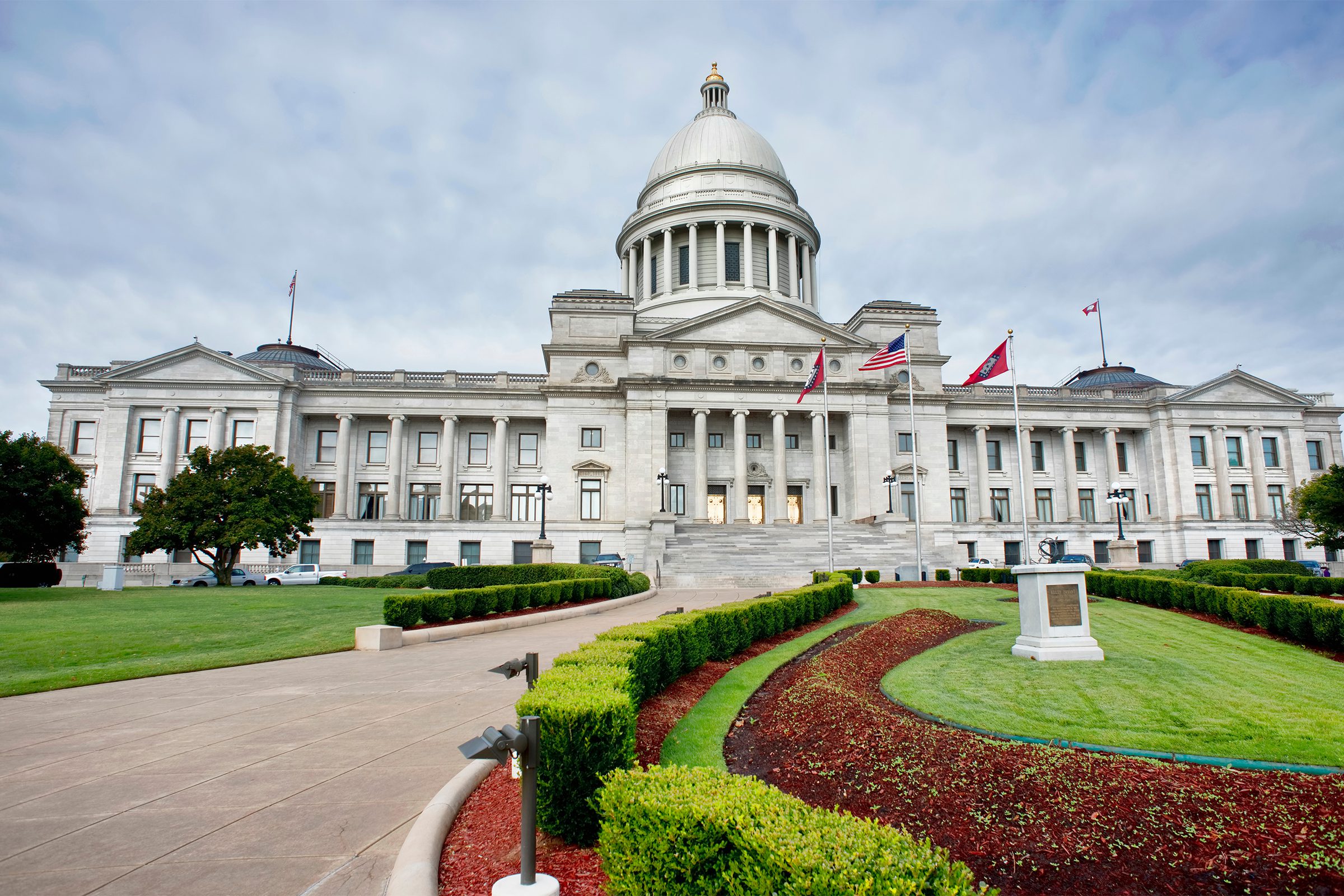The image shows the front view of a large white government building with a dome, columns, and several flags flying, surrounded by manicured lawns and landscaping.