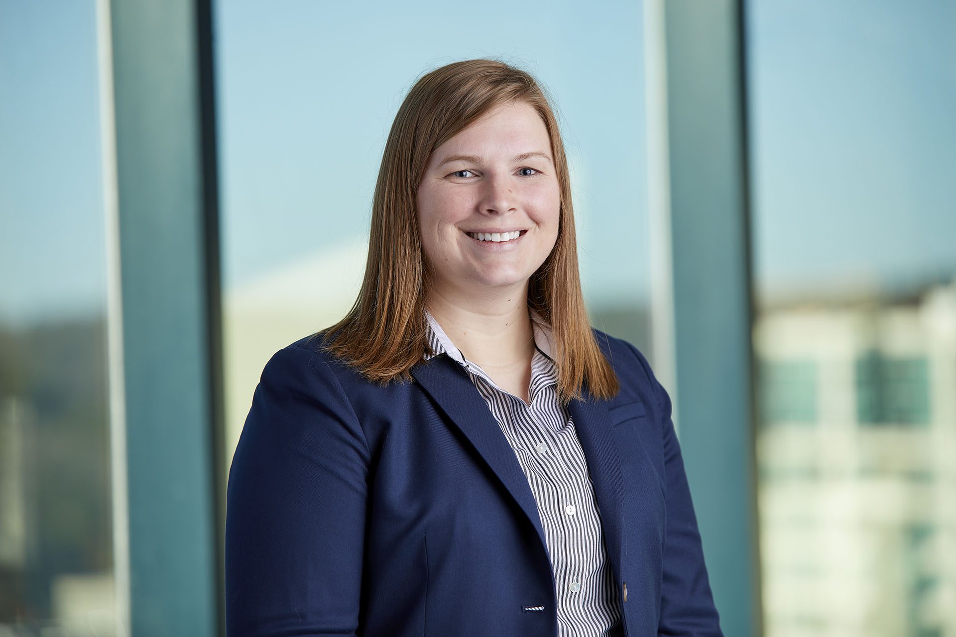 A woman in a navy blazer and striped shirt stands indoors in front of large windows, smiling at the camera.