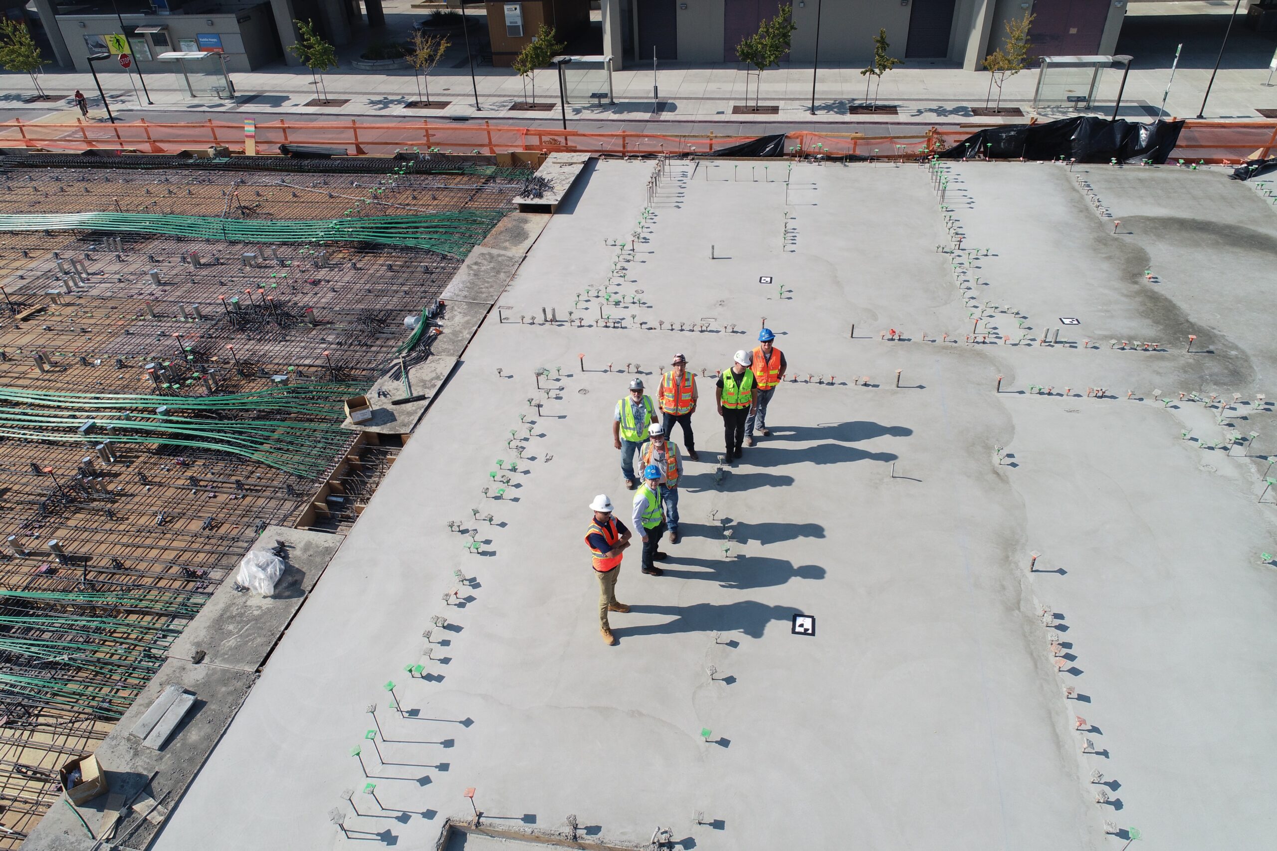 A group of six construction workers in safety gear stand on a large concrete slab at a building site, with materials and rebar visible nearby.