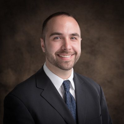 A man in a suit and tie smiles at the camera against a brown studio backdrop.