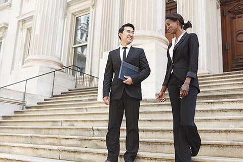 Two business professionals in suits walk and talk on the steps outside a large, columned building.