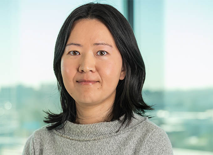 A woman with straight, shoulder-length black hair wearing a grey sweater stands in front of a window with an outdoor background.
